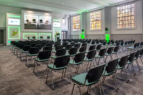 Lots of chairs laid out in a large conference style hall with guest speaker chairs on a plinth that are also projected onto a large screen above.
